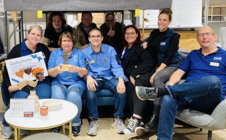 Neun Menschen, vier Frauen und fünf Männer, sitzen auf einem Sofa. Eine Frau hält einen Pokal in der Hand, eine andere einen Gutschein. 