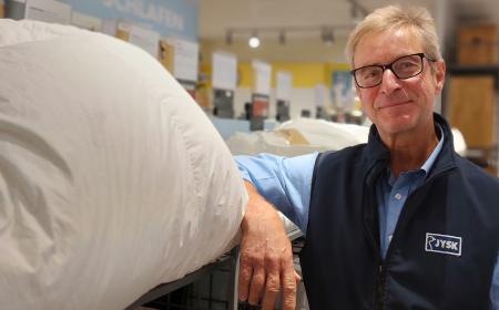 Ein Mann mit Brille und kurzen Haaren lächelt in die Kamera. Er lehnt lässig an einem Regal, aus dem ein Kissen rausschaut.