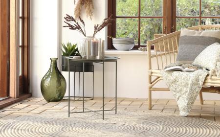 Eine Bank in Bambusoptik mit Kissen und Decken sowie ein Beistelltisch mit einer Vase und Kerze. Im Hintergrund sieht man eine Fensterfront sowei weitere Wohnaccessoires