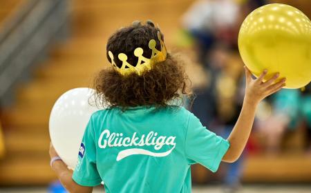 Ein Mädchen mit lockigen Haaren und einer Pappkrone auf dem Kopf von hinten. Auf ihrem Shirt steht "Glücksliga". In den Händen hällt sie zwei Luftballons. 