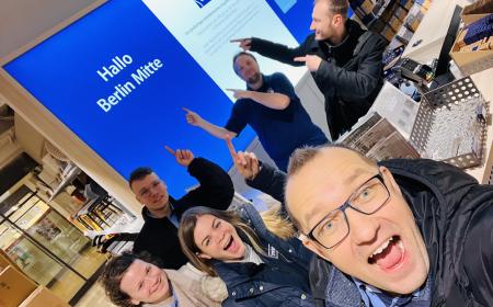 Sechs Menschen – zwei Frauen und vier Männer – stehen an einem Kassentresen. Sie zeigen auf die Aufschrift "Hallo Berlin Mitte"
