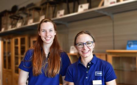 Zwei Frauen – eine mit langen Haaren und eine mit Pferdeschwanz und Brille – lächeln in die Kamera. Beide tragen JYSK-Poloshirts 