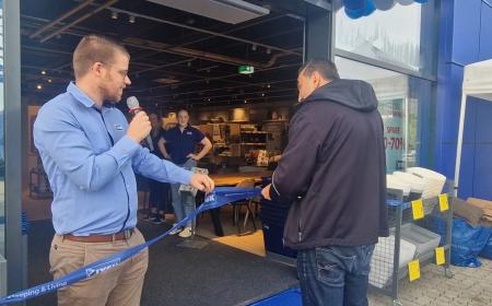 Zwei Männer stehen vor einem JYSK Store. Einer hält in der einen Hand ein Mikrofon und in der anderen ein dickeres Band. Der zweite Mann hält eine Schere in der Hand. 