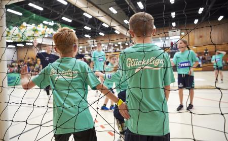 Zwei jungen stehen in einem Handballtor. Sie stehen mit dem Rücjen zur Kamera. Sie tragen Shirts mit der Aufschrift Glücksliga. im Hintergrund stehen weitere Kinders in Trikots. 