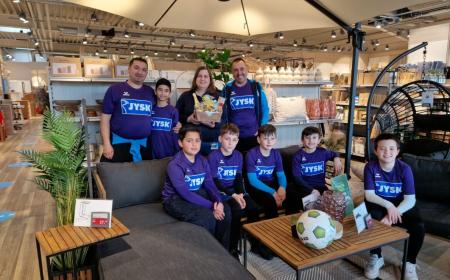 Mehrere Kinder und drei Erwachsene sitzen und stehen in einem JYSK-Store. Eine Frau hält einen kleinen Geschenkkorb in der Hand 