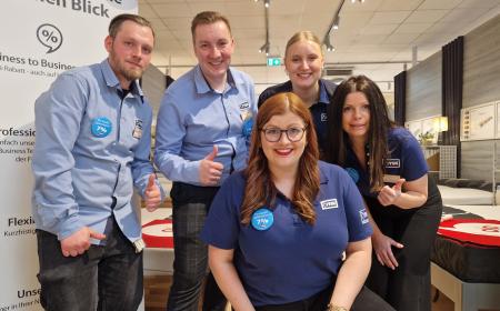 Fünf Menschen – drei Frauen und zwei Männer – tragen JYSK-Hemden und JYSK-Poloshirts. Sie lächeln in eine Kamera