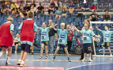 Kinder in Handballtrikots stehen vor einem Tor. Sie halten die Arme nach oben, um einen Ball abzuwehren. 