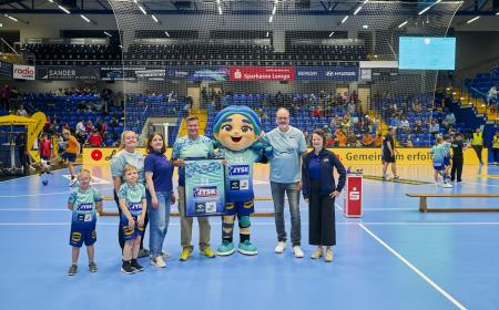 Sieben Menschen und ein Maskottchen stehen auf einem Handballfeld
